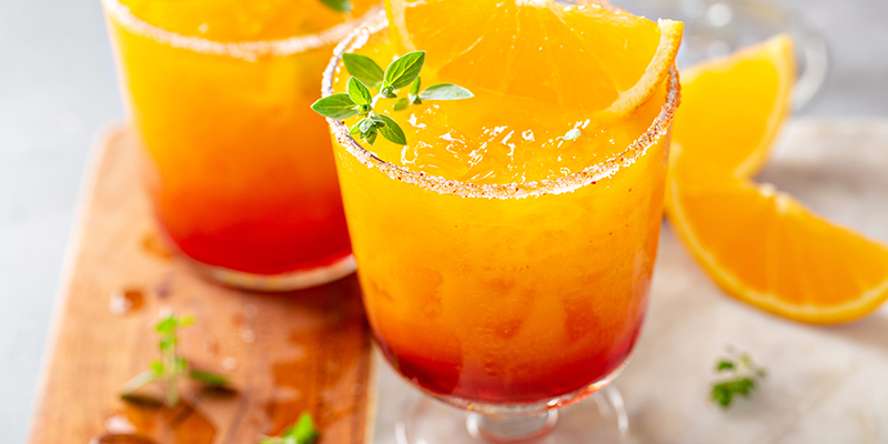 Jugos cítricos adornados con gajos de naranja sobre una superficie blanca. Los acompaña una tabla de madera.