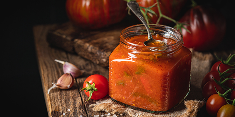 Salsa de tomate en un recipiente de vidrio sobre una mesa de madera, acompañado de varios tomates y ajos.