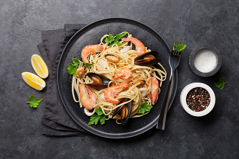 Vista superior de un plato de pasta con mariscos, almejas y especias con un tenedor en un plato negro. Alrededor hay una servilleta, semillas, sal y gajos de limón. Todo sobre un fondo oscuro.