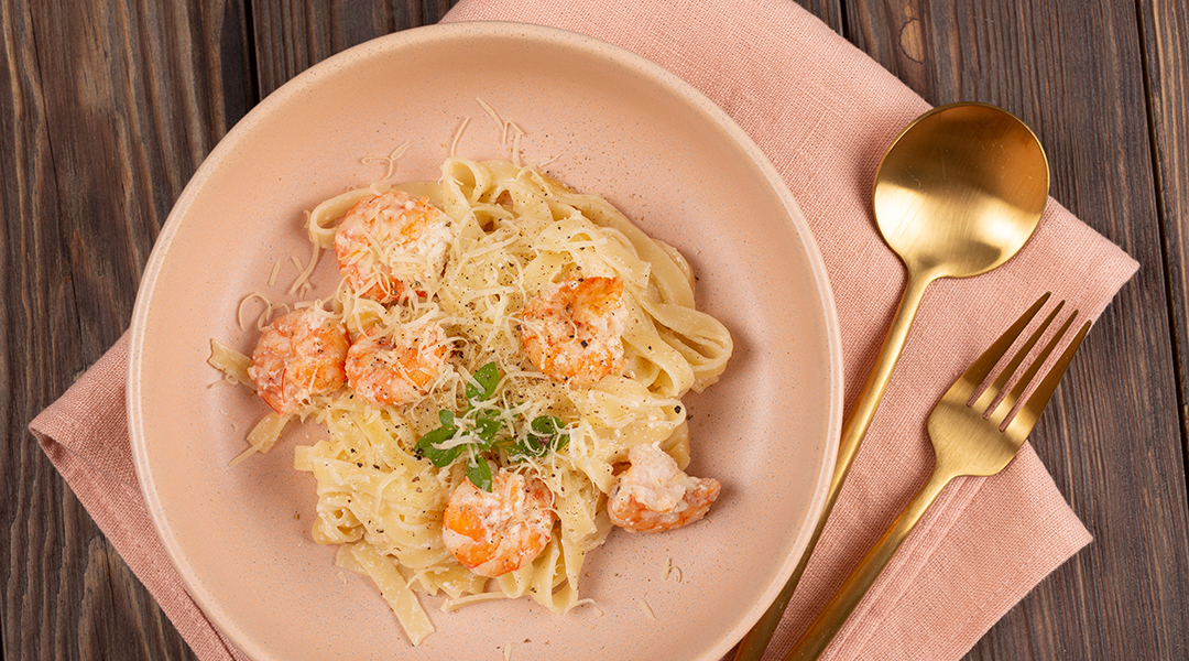 Plato de color rosa pastel con fettuccine, camarones, queso parmesano y perejil, acompañado de una servilleta de tela y cubiertos dorados. Todo sobre una mesa de madera.