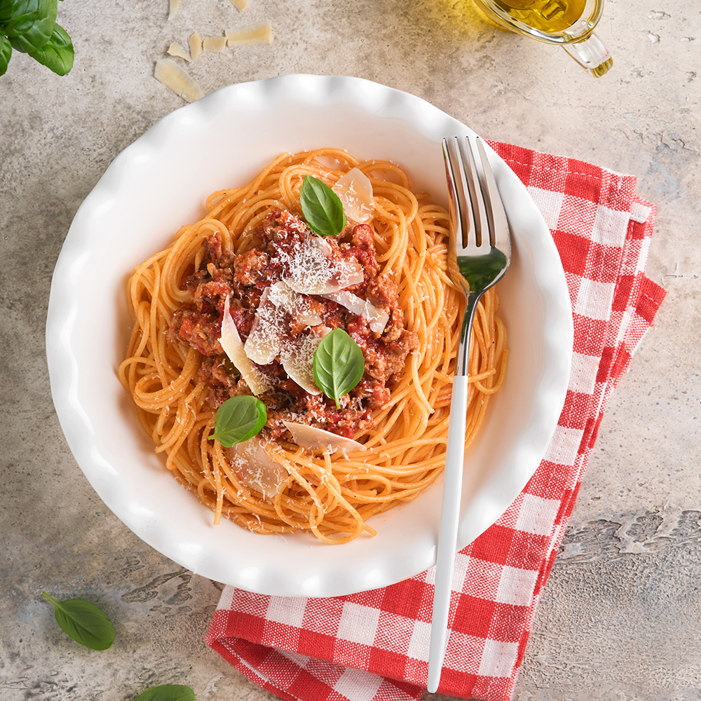Vista superior de un plato de linguine con carne, queso parmesano y algunas hojas de albahaca junto con un tenedor. Lo acompaña una servilleta de tela de cuadros rojos y blancos, un recipiente de vidrio con aeceite de oliva y más hojas de albahaca. Todo sobre un fondo gris.