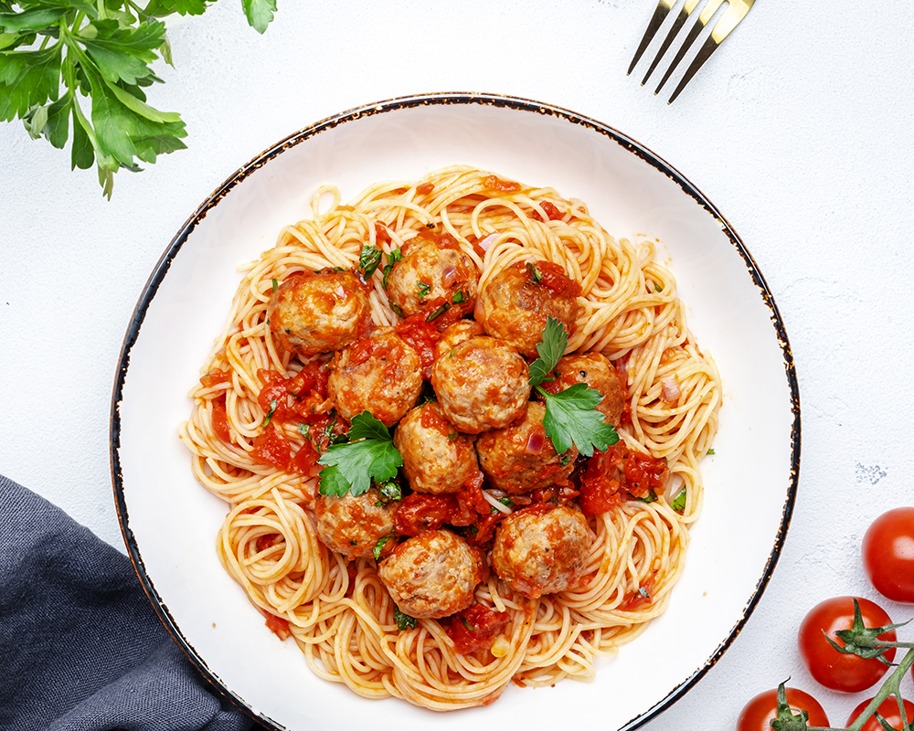 Vista superior de un plato de vermicelli con camarones, especias y pasta de tomate sobre una superfice blanca. Lo acompañan dos tenerdores, sal, primienta, tomates y hojas verdes de cilantro.