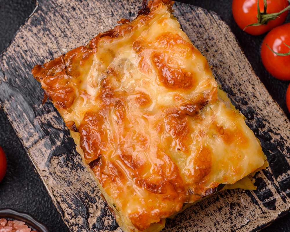 Vista superior de una porción de lasagna sobre una tabla de madera. La acompañan pequeños recipientes con especias.