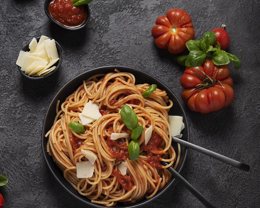 Vista superior de un plato de spaghetti con salsa de tomate, albahaca y queso. Alrededor hay tomates. Todo sobre un fondo oscuro.