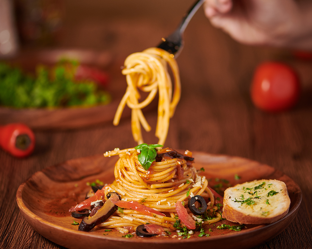 Vista de un plato oscuro de spaghetti con carne y verduras, acompañado de una rodaja de pan con especias. Al fondo se aprecia un tomate y algunas hojas verdes; todo sobre una superficie de madera.