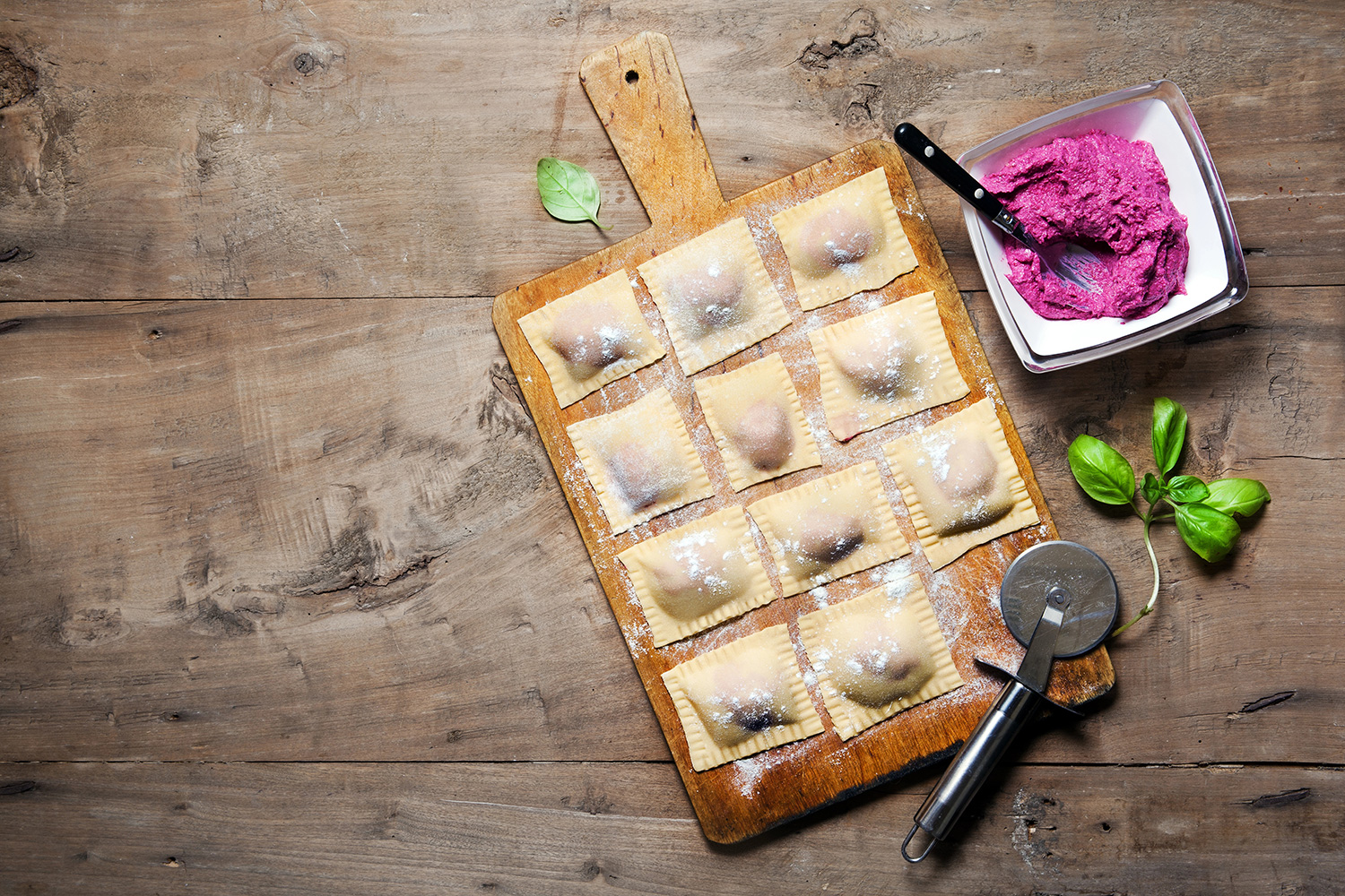 Vista superior de raviolis rellenos con mezcla de arándanos y queso sobre una tabla de madera. Los acompaña un cortador de pasra, hojas frescas de menta y un bowl pequeño con el relleno; todo sobre una mesa de madera