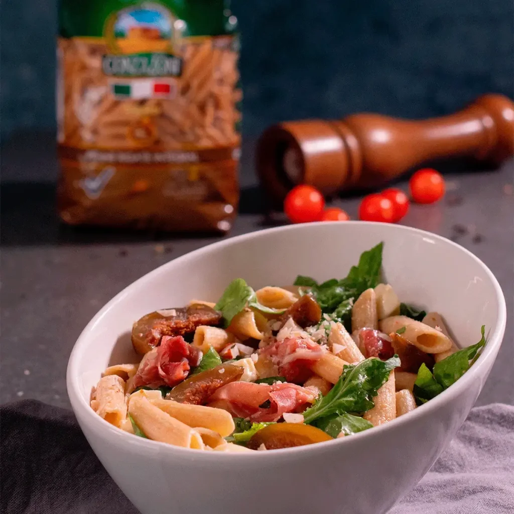 Ensalada de pasta Conzazoni con jamón, tomates cherry, rúcula y queso rallado, servida en un bol blanco.