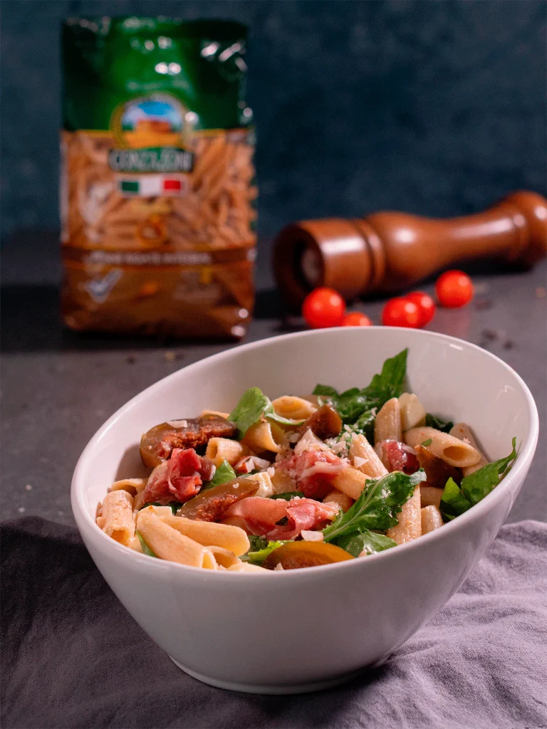 Ensalada de pasta Conzazoni con jamón, tomates cherry, rúcula y queso rallado, servida en un bol blanco.