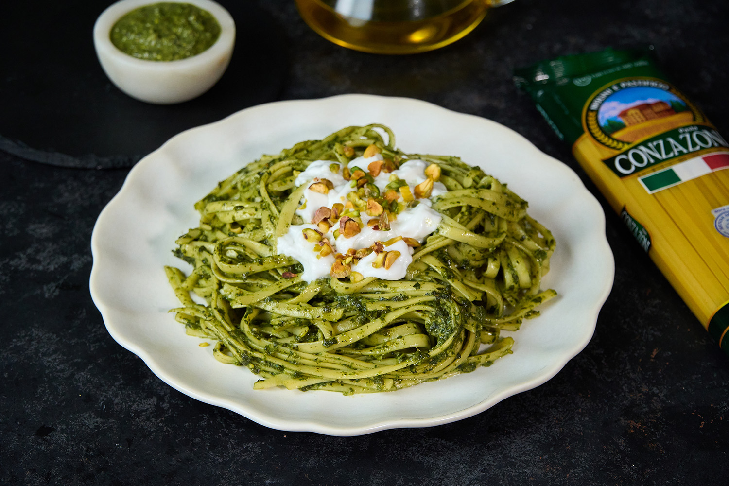 Vista de un plato con fettuccine con salsa de pesto de pistacho y burrata. Alrededor hay un paquete de Fettuccine Conzazoni, un bowl pequeño con salsa de pesto. Todo sobre un fondo oscuro.