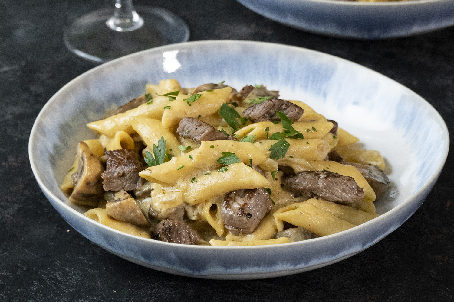Vista de dos platos de penne rigate con stroganoff y especias. Al fondo hay copas de vino, un bowl con salsa verde. Todo sobre un fondo oscuro.