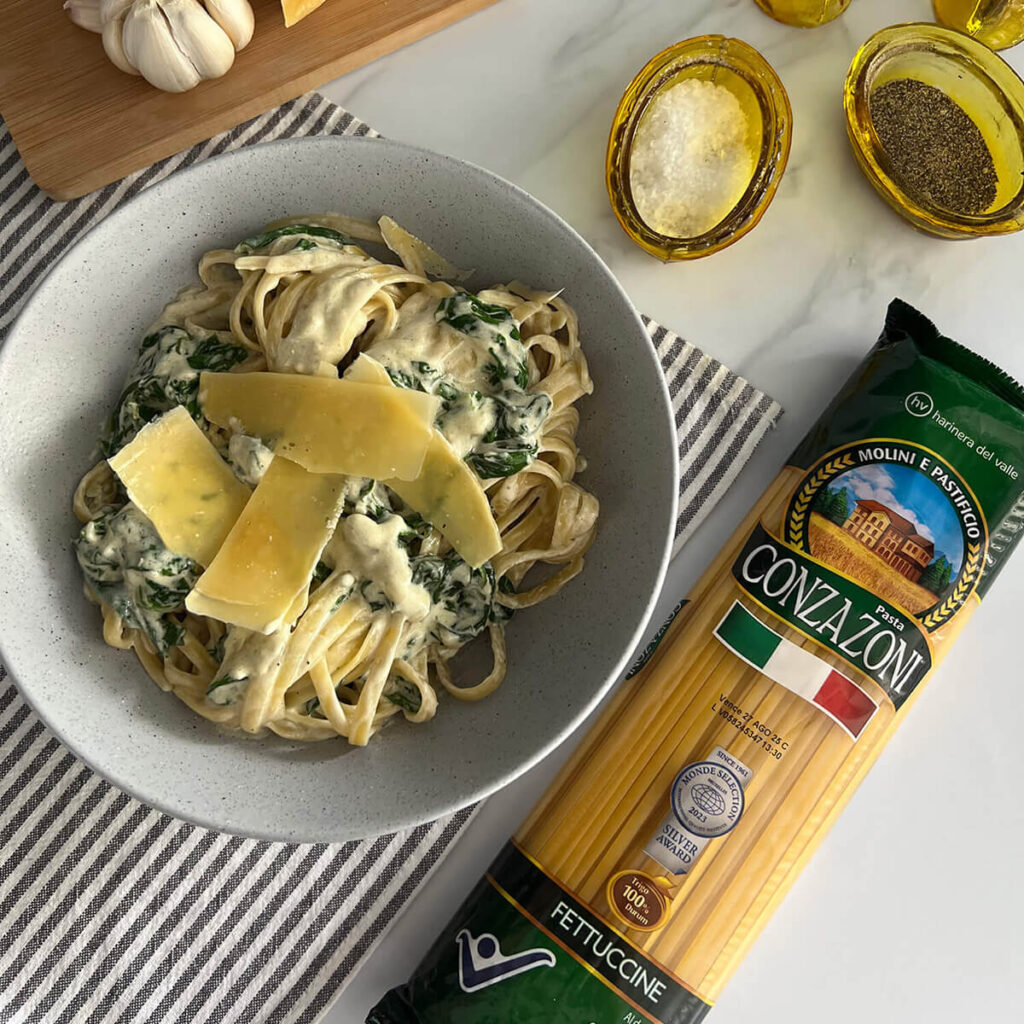 Vista superior de un plato de fettuccine con espinacas y queso azul acompañado de un empaque de Fettuccine de Conzazoni de sal, pimienta, ajos y queso azul. Todo sobre un fondo claro