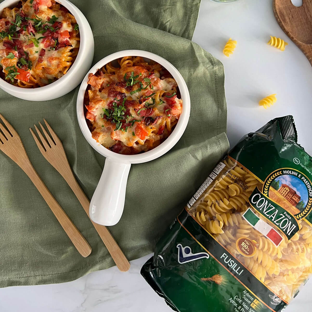 Vista superior de dos bowls de fusilli gratinado con salami, tomates y queso. Alrededor hay un empaque de Fusilli de Conzazoni y cubiertos de mandera. Todo sobre un fondo claro