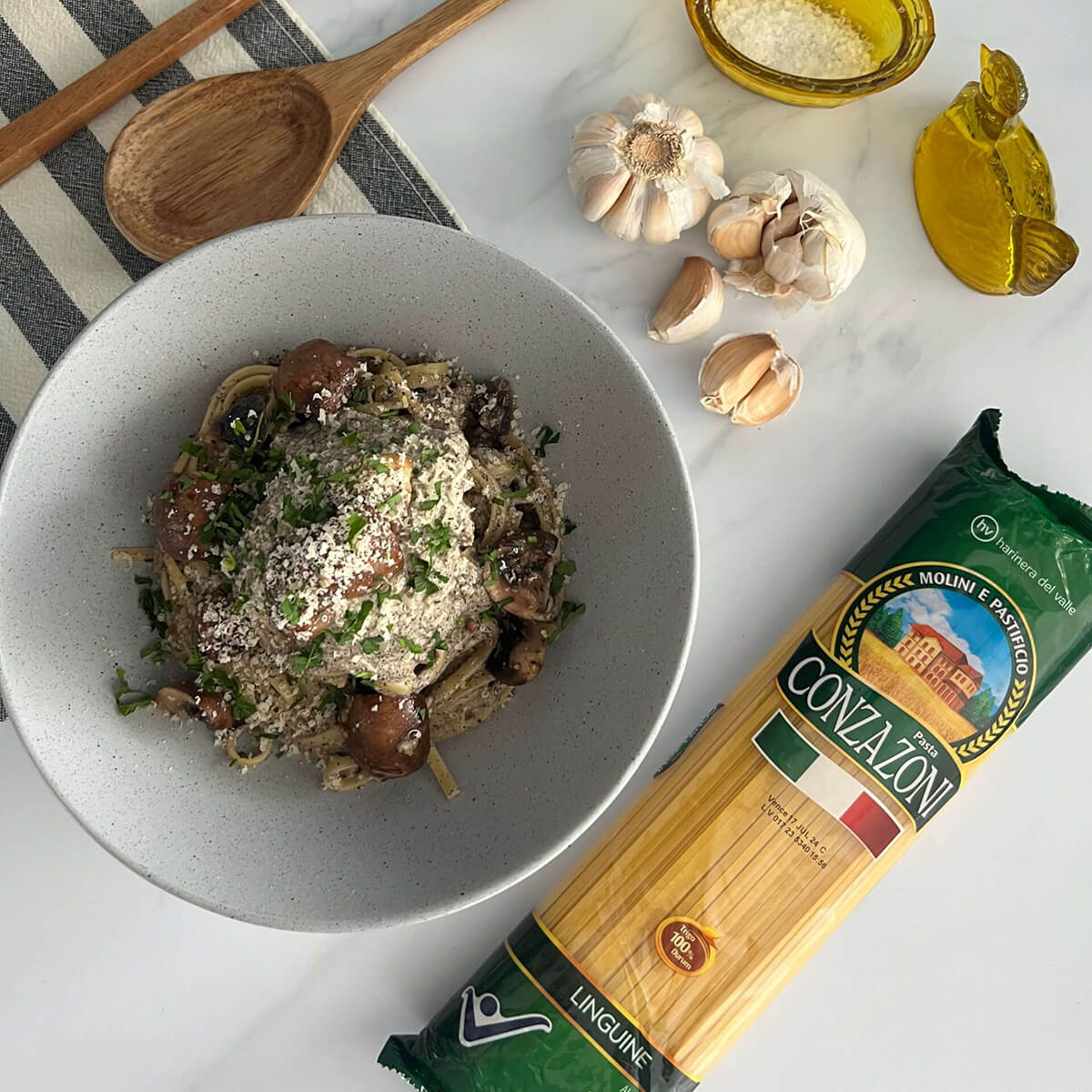 Vista superior de un plato de linguine con crema de hongos, acompañado de utensilios de cocina, ajos y sal. Todo sobre un fondo claro