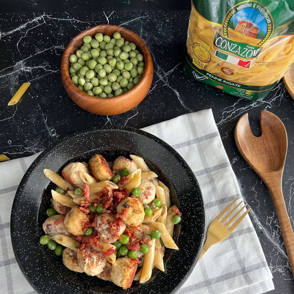 Vista superior de un plato de penne rigate con albóndigas de pollo acompañado de un tendeor, un bowl con arvejas verdes, una cuchara de madera y un empaque de Penne Rigate Cozazoni. Todo sobre un fondo oscuro