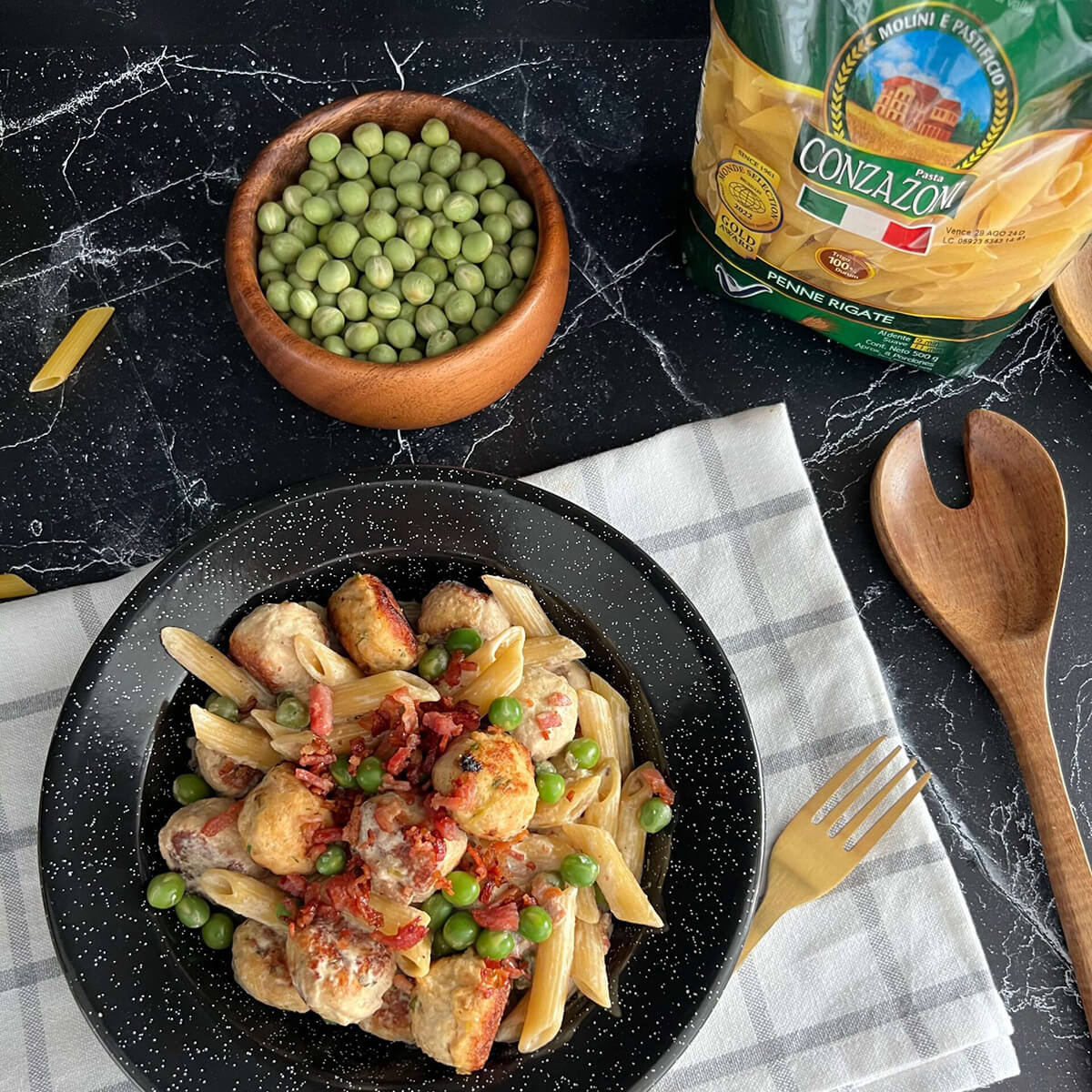 Vista superior de un plato de penne rigate con albóndigas de pollo acompañado de un tendeor, un bowl con arvejas verdes, una cuchara de madera y un empaque de Penne Rigate Cozazoni. Todo sobre un fondo oscuro