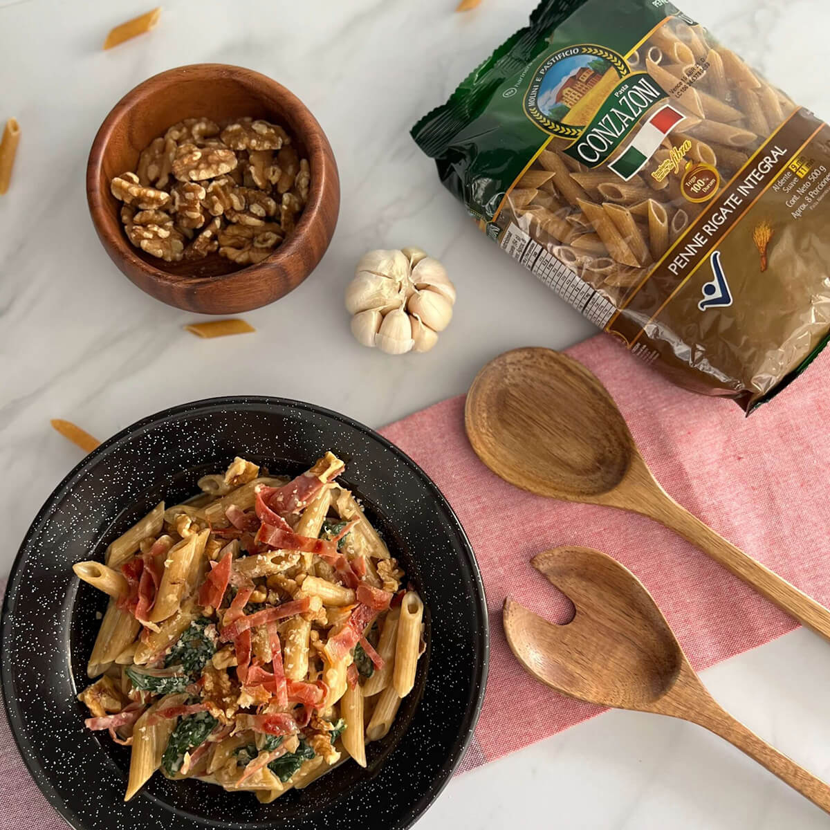 Vista superior de un plato de penne integral con espinacas a la crema y jamón serrano. Alrededor hay un empaque de Penne Rigate Integral de Conzazoni, utensilios de cocina y dientes de ajo sobre una servilleta de tela. Todo en una superficie cubierta con un mantel blanco