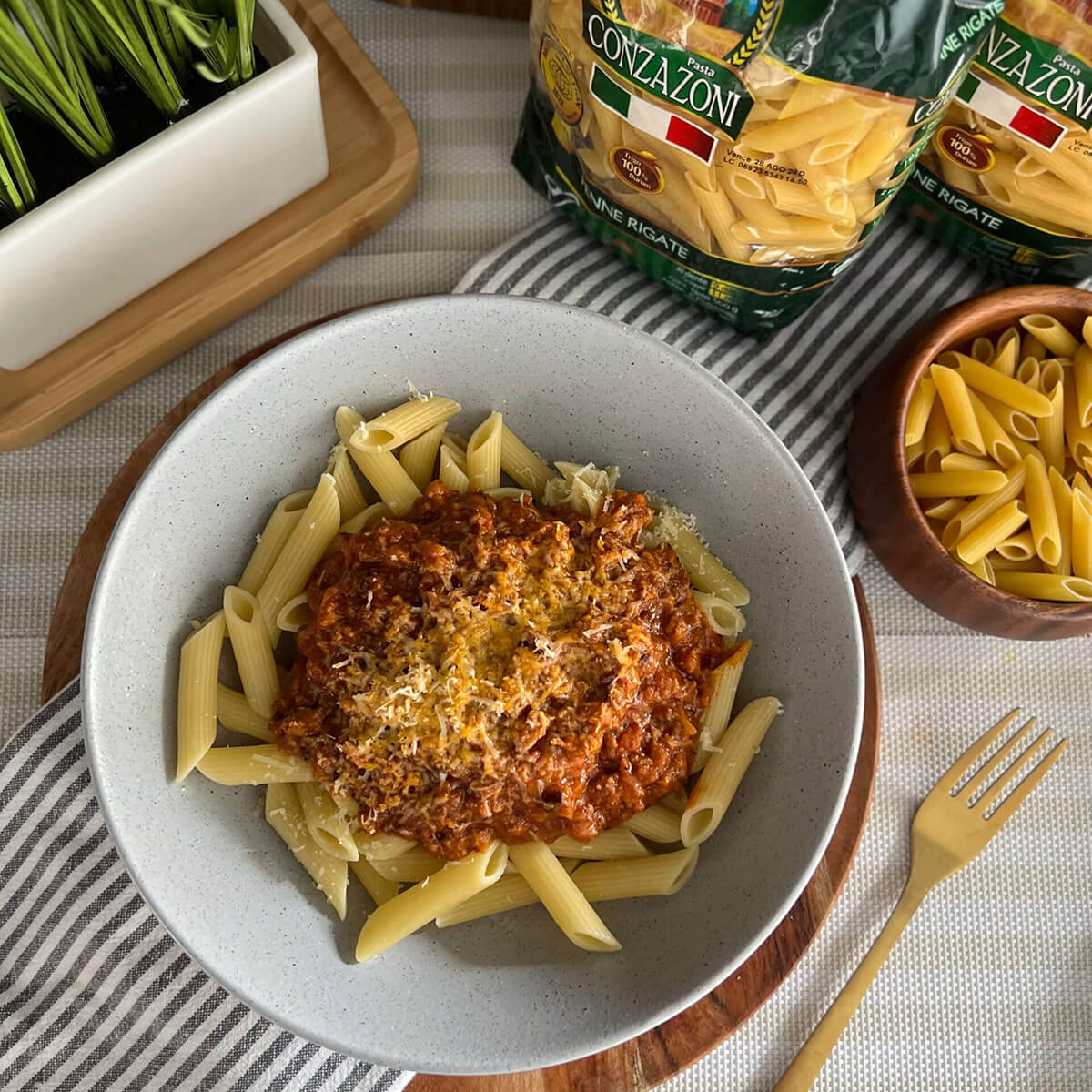 Vista superior de un plato de penne rigate con ragú de cerdo y crema. Alrededor hay un tenedor, empaques de Penne Rigate de Conzazoni y un bowl con pasta cruda. Todo sobre una mesa con mantel