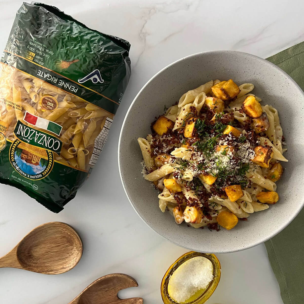 Vista superior de un plato de penne rigate con suero, tocino y maduro. Acompañado de utenislios de cocina y un empaque de Penne Rigate de Conzazoni. Todo sobre un fondo claro.