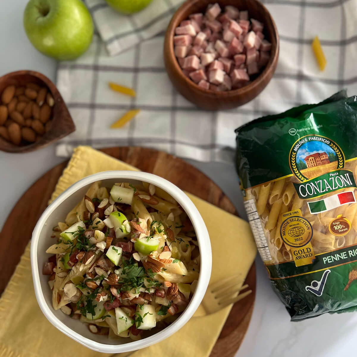Vista superior de un plato de penne rigate con tocineta, manzana y almendras sobre una servilleta de tela. Alrededor hay un empaque de Penne Rigate de Conzazoni, un bowl con tocineta en cubos y manzanas.
