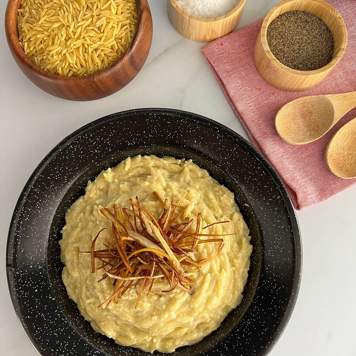 Vista superior de un plato de un plato de risoni con crema de puerros. Alrededor hay un bowl con risoni crudo, sal y pimienta. Todo sobre un fondo claro