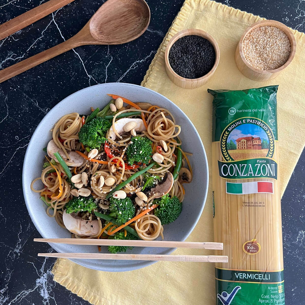 Vista superior de un plato de vermicelli asiático con vegetales acompañado de palillos chinos, un empaque de Vermicelli Conzazoni y utensilios de cocina. Todo sobre una superficie de mármol oscuro.