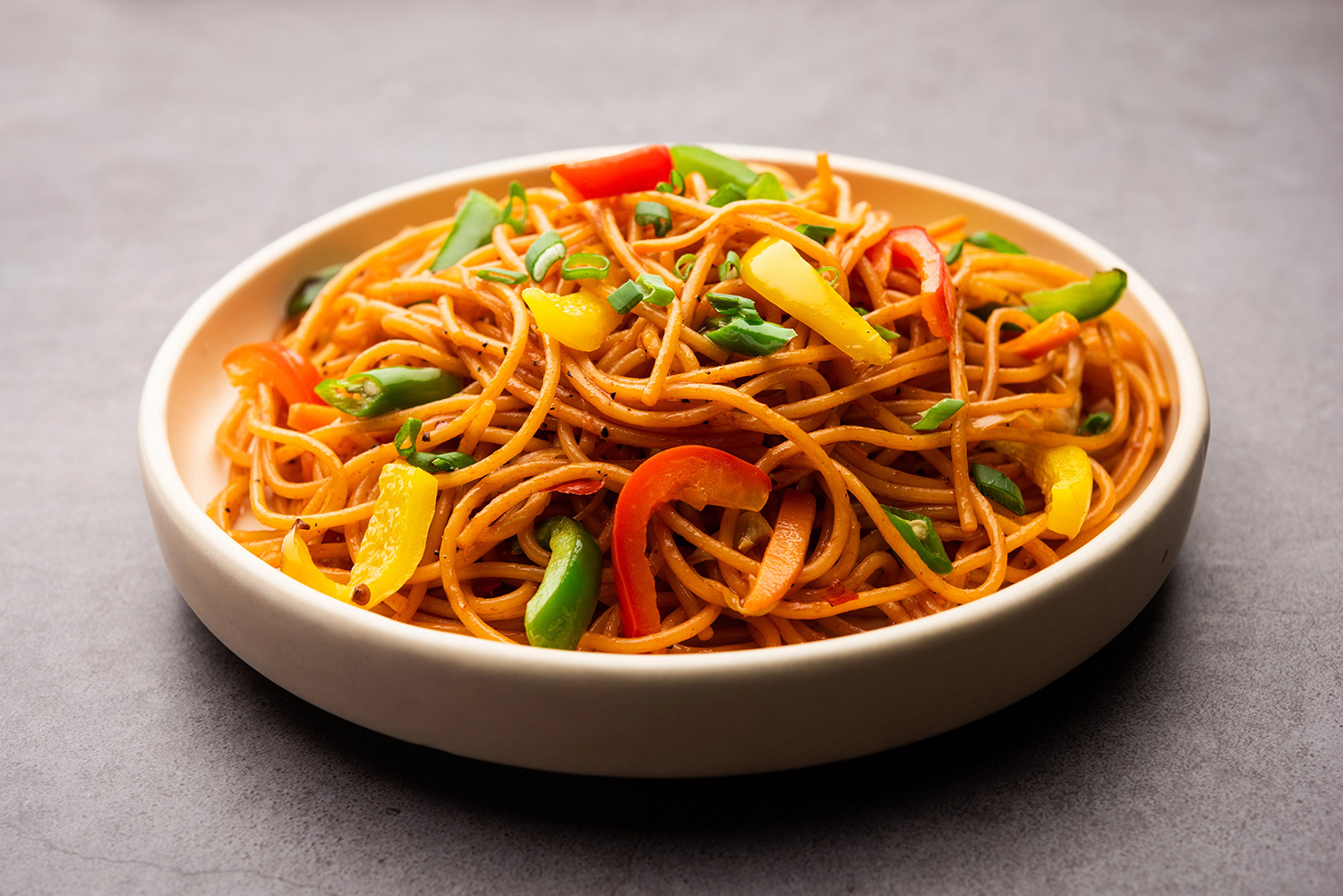 Vista superior de un plato de spaghetti con verduras saltadas como calabacín, pimentón rojo, amarillo y verde con cebollín en trocitos, sobre una superficie gris.