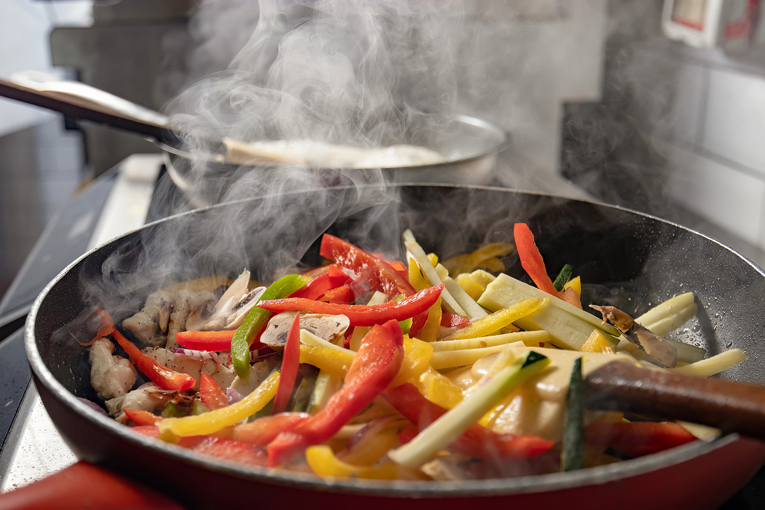 Vista de una sartén caliente con trozos de diferentes verduras picadas en julianas, como pimetón amarilo, y rojo, y calabacín en un ambiente de cocina.