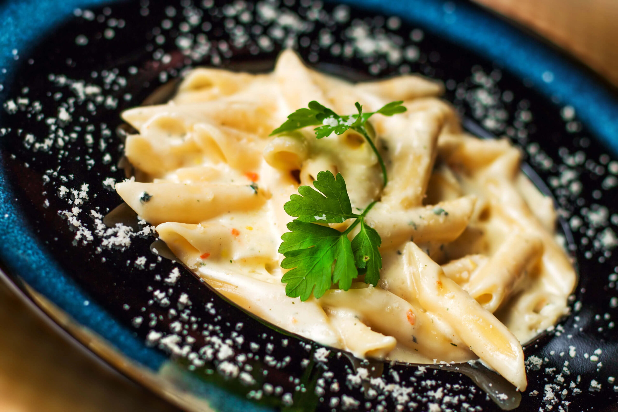 Vista superior de un plato de penne rigate con cuatro quesos fundidos, perejil decorativo, sobre un plato negro con queso parmesano espolvoreado