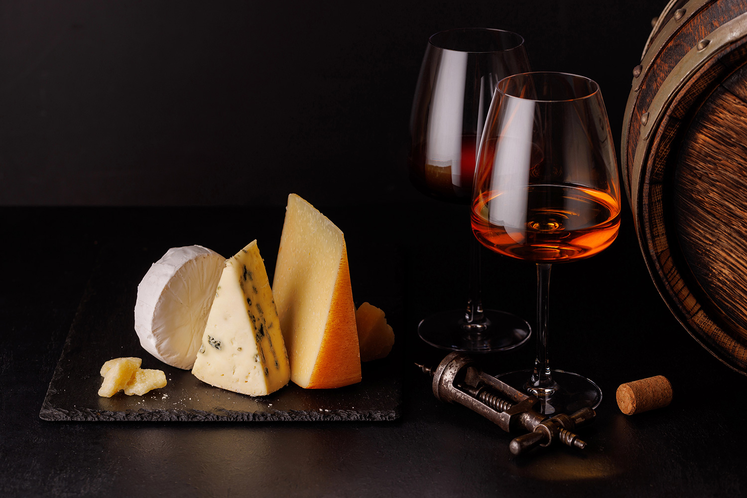 tabla cuatro quesos, copas de vino, barril de conserva, sobre mesón negro