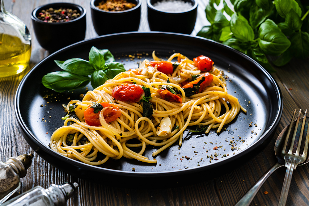 Visual de pasta puttanesca servida en un plato negro junto a 3 tarros de ingredientes para preparación.