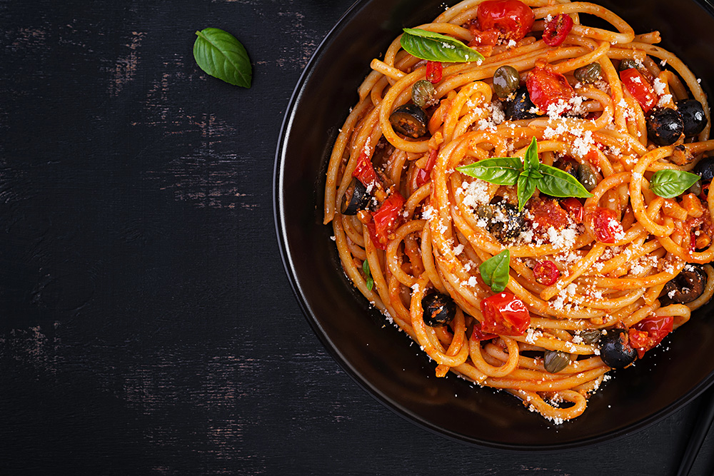 Media visual de pasta puttanesca servida en un plato negro sobre una superficie oscura.