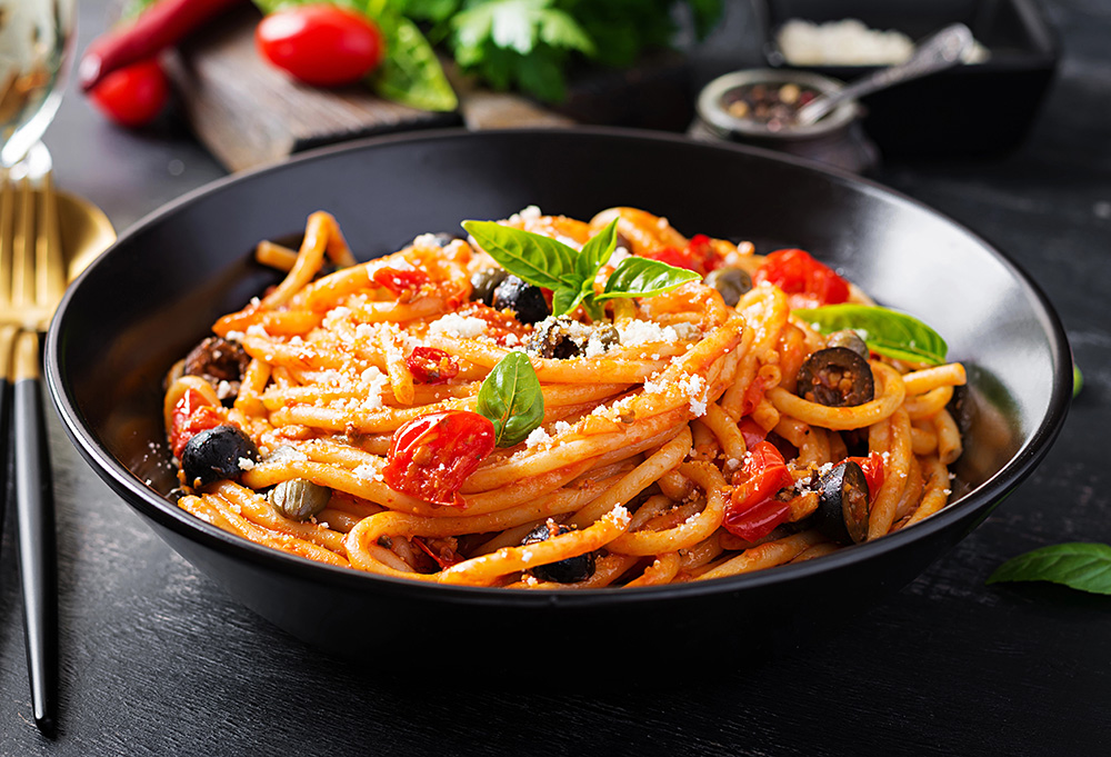 Visual de pasta puttanesca servida en un plato negro con acompañantes como tomates, hojas verdes y cubiertos