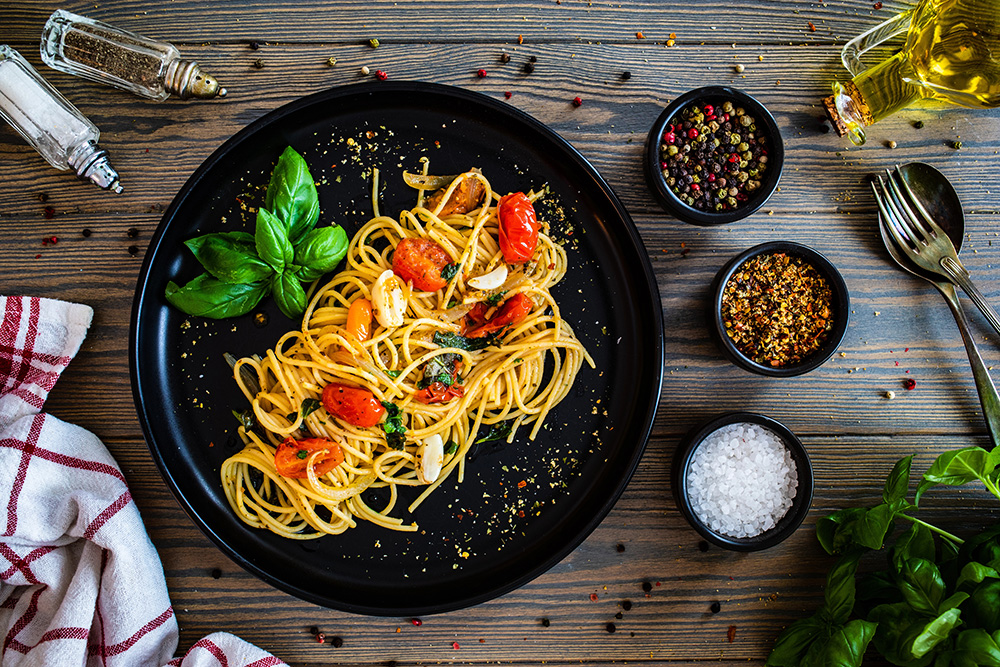 Visual de pasta puttanesca servida en un sartén negro sobre una mesa amaderada, con cubiertos y recipientes con especias que la acompañan.