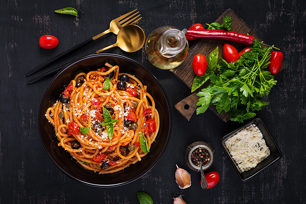 Visual de pasta puttanesca servida en un plato negro con ingredientes y cubiertos sobre la mesa.