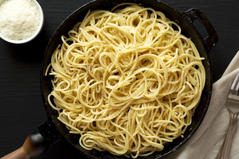Pasta cacio e pepe sobre olla negra en un fondo oscuro y queso en un recipiente como decoración