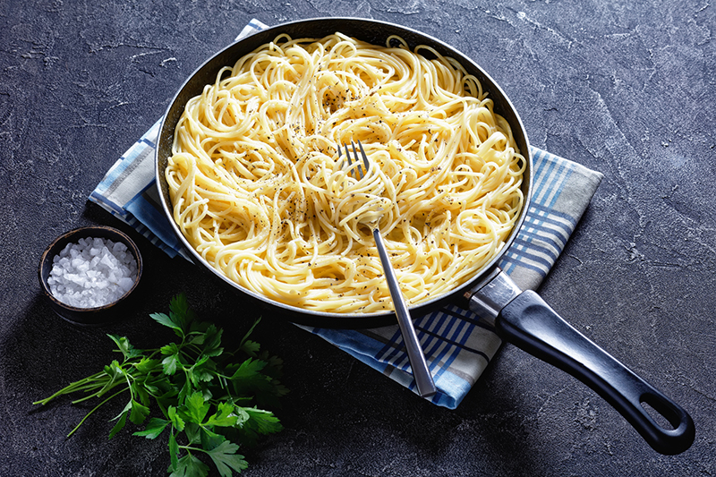 Pasta cacio e pepe en una sartén negra sobre una toalla de cocina junto con sal en un repipiente y unas hojas de cilantro para decorar la mesa