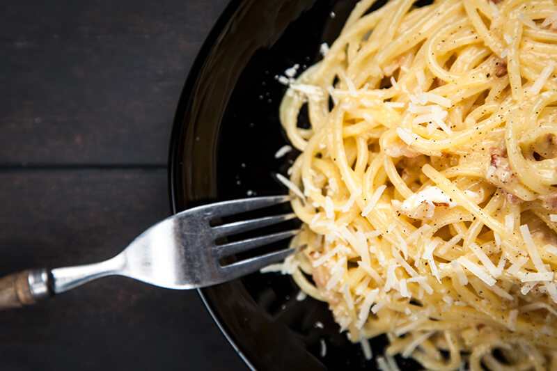 Pasta cacio e pepe en un plato negro junto con un tenedor, todo sobre una superficie oscura