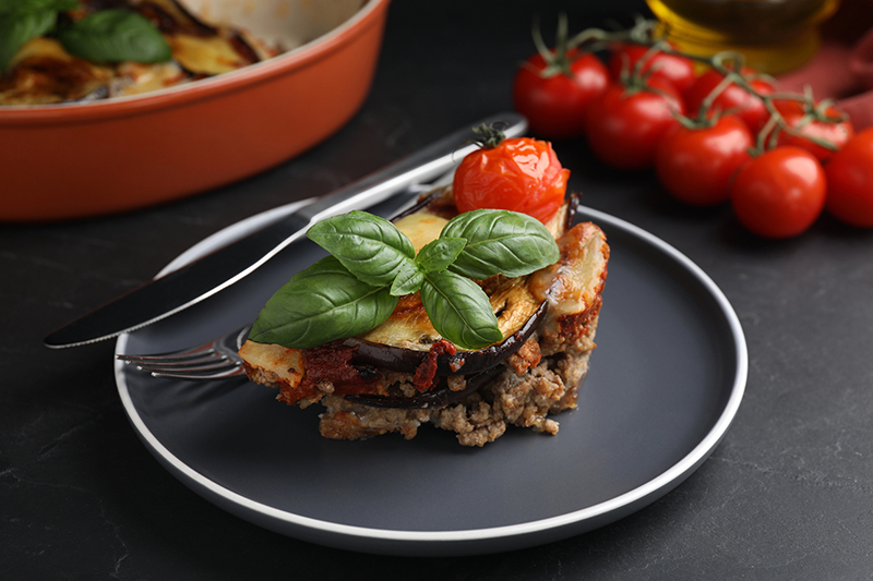 Visual de lasagna de berenjena decorada con hojas de albahaca y tomate, servida en un plato y mesa color negro. Alrededor hay tomates de decoración