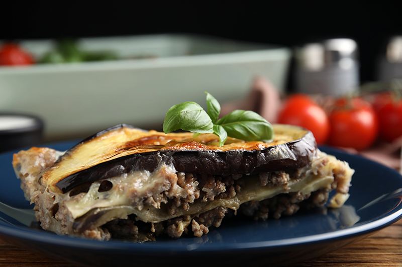 Porción de lasagna de berenjena decorada con hojas de albahaca, servida en un plato azul sobre una mesa de madera