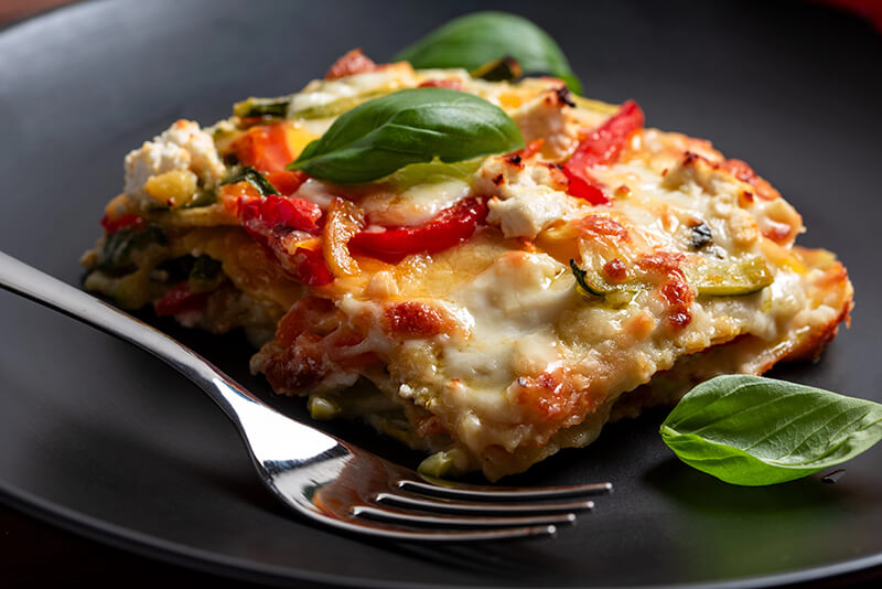 Porción de Lasagna de berenjena decorada con hojas de albahaca, sobre un plato negro redondo