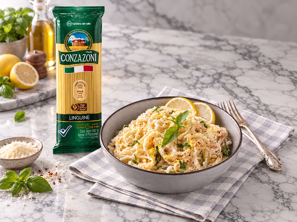 Plato hondo con linguine en salsa cremosa de limón y parmesano, decorado con hojas frescas de albahaca, ralladura de limón y un toque de especias, acompañado de rodajas de limón y cubiertos metálicos sobre una servilleta de cuadros grises, junto al empaque de Linguine Conzazoni. Todo es una superficie clara de mármol.
