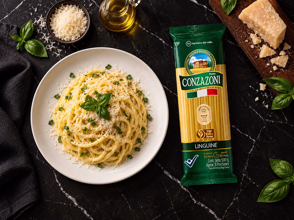 Plato de linguine con limón y parmesano, decorado con hojas frescas de albahaca y acompañado de trozos de queso parmesano y una servilleta negra de tela, junto al empaque del producto Linguine Conzazoni. Todo sobre una superficie oscura de mármol.